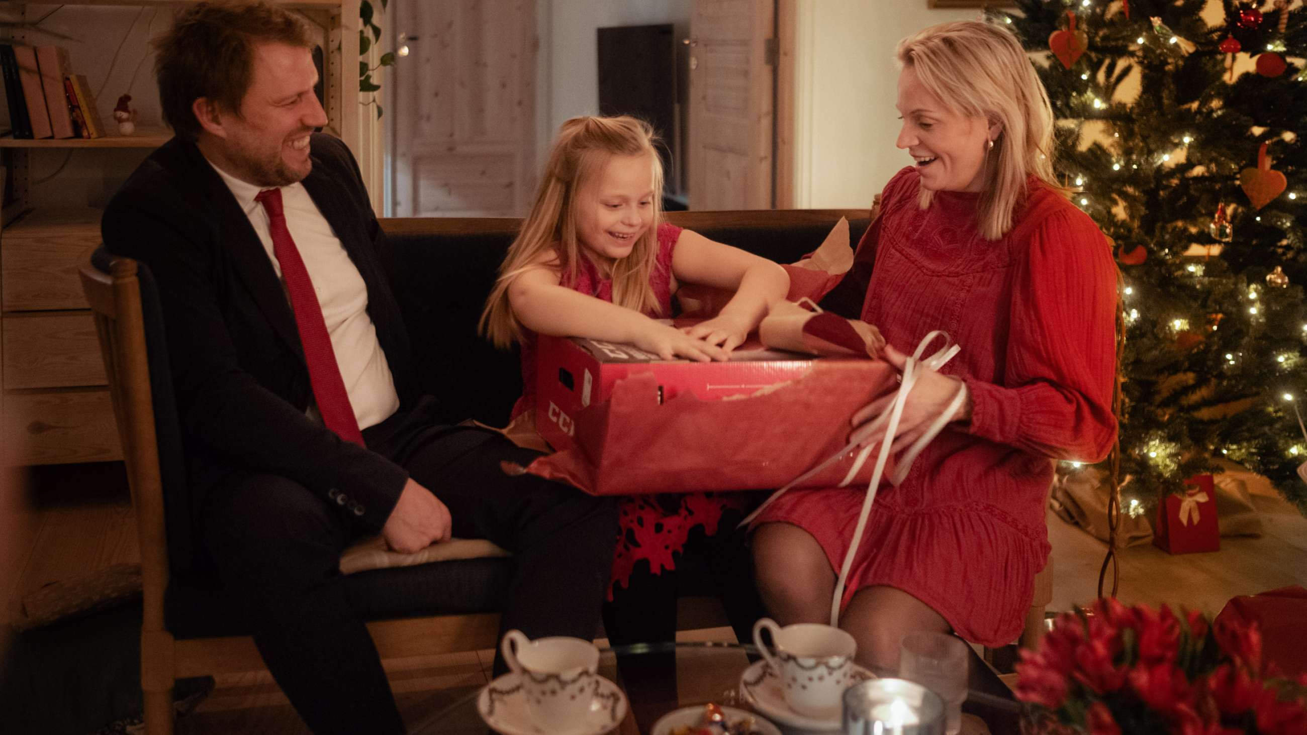 Et barn sidder mellem en mand og en kvinde i en sofa juleaften og åbner en julegave