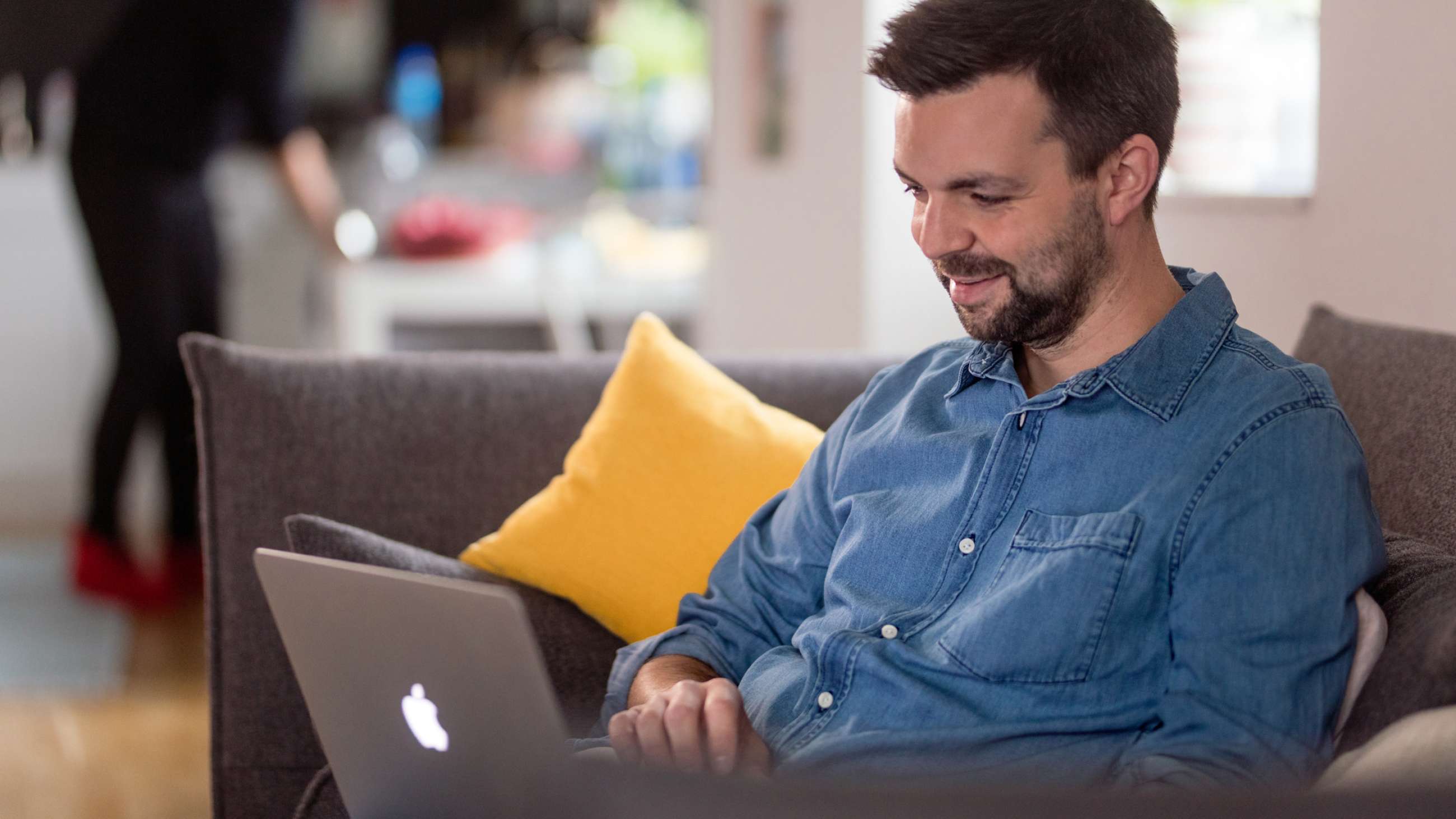 Ung mand med skæg og jeansskjorte sidder smilende i sofaen med en laptop i skødet.