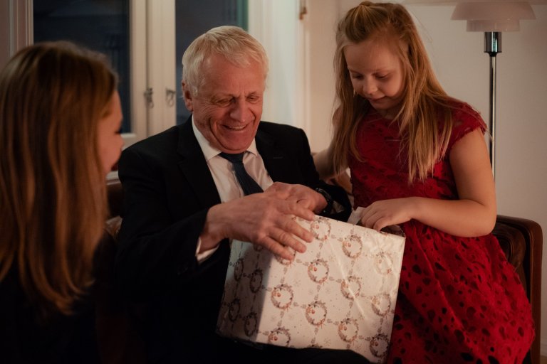 En ældre mand sidder i en sofa med sit barnebarn ved siden af ​​og åbner en julegave