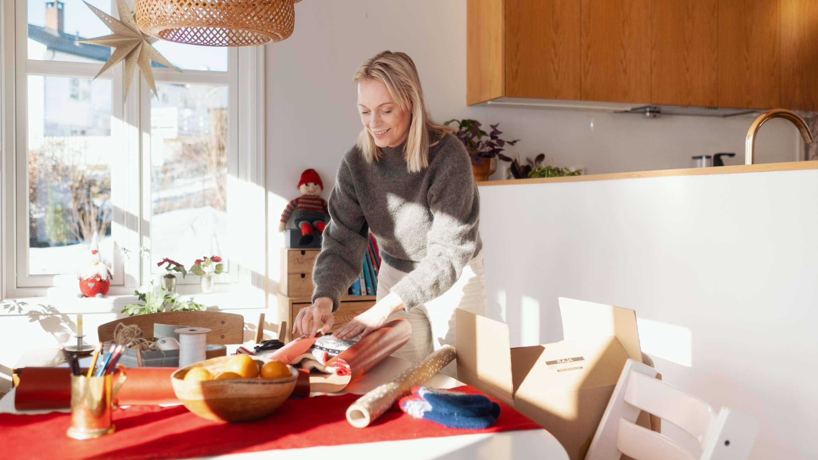 En kvinde læner sig smilende over et bord og pakker en sweater i julegave.