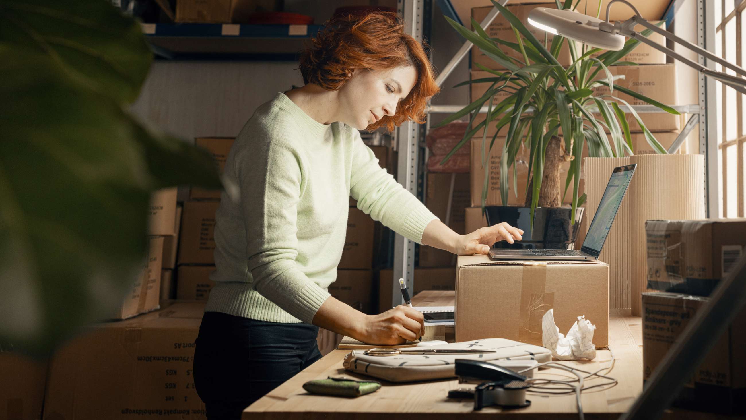 Kvindelig indehaver af en netbutik står med en papkasse på et arbejdsbord med mange kasser i baggrunden. Hun skriver på en blok, mens hun holder den anden hånd på en åben laptop.
