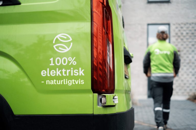 En el-varebil fra Bring som er parkeret foran et hus, mens chaufføren er på vej mod hoveddøren for at leverer en større pakke