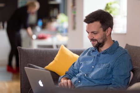 Ung mand med skæg og jeansskjorte sidder smilende i sofaen med en laptop i skødet.