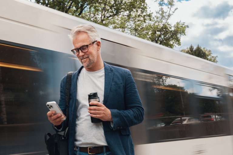 Mand står ved en togstation og ser på sin mobiltelefon.